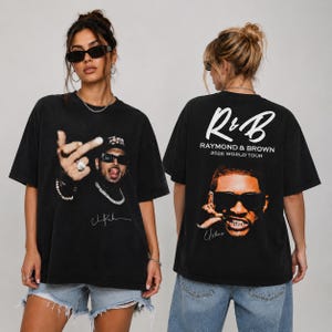 Może przedstawiać: Czarny t-shirt z krótkim rękawem z nadrukiem graficznym przedstawiającym osobę w okularach przeciwsłonecznych i biżuterii. Przód pokazuje gestykulującą osobę, a tył zawiera napis R&B RAYMOND & BROWN 2026 WORLD TOUR nad portretem osoby.