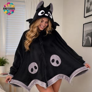 Op de afbeelding: Een zwarte cape met capuchon en een op een wezen geïnspireerd ontwerp met grote witte ogen op de capuchon, kleine zwarte vleermuisachtige vleugels aan de zijkanten en twee cirkelvormige grijze patches aan de voorzijde. De zoom is afgewerkt met een brede, lichtgrijze rand.