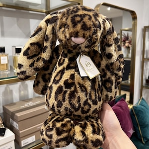 May include: A plush stuffed animal in the shape of a rabbit with long ears, covered in a tan and dark brown leopard print pattern. The toy features a soft pink nose and a white paper tag attached to its neck with the text AIRAHI printed on it.