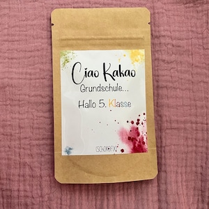 Könnte beinhalten: Ein rechteckiger Beutel aus braunem Kraftpapier mit einem weißen Etikett, das bunte Farbspritzer aufweist. Der Text lautet Ciao Kakao Grundschule... Hallo 5. Klasse und enthält unten den Markennamen Schokofix in kleiner schwarzer Schrift.