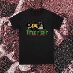 Pode incluir: Uma t-shirt preta de manga curta com um gráfico de um leão amarelo sobre blocos verdes. O texto "TITLE FIGHT" aparece em letras verdes acima do texto vermelho "KINGSTON PA USA" centrado na parte frontal da peça de vestuário.