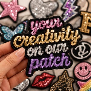 Pode incluir: Um emblema de tecido preto com o texto your creativity on our patch bordado em lantejoulas. A palavra your é rosa, creativity é dourada, on our é prateada e patch é roxa. Outros emblemas de lantejoulas variados estão visíveis ao fundo.
