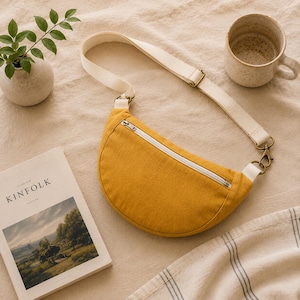 May include: A crescent-shaped mustard yellow crossbody bag featuring a white zip closure and an adjustable off-white canvas strap with brass-toned metal hardware clips. The bag has a smooth fabric texture and a curved bottom edge design.