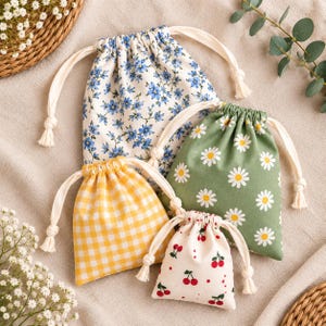 Puede incluir: Cuatro bolsas de tela con cordón de diferentes tamaños. Una presenta un patrón floral azul, otra un estampado de margaritas verdes, otra un cuadro vichy amarillo y blanco, y la más pequeña muestra un fondo blanco con un patrón de cerezas rojas.