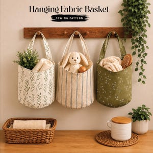 Peut inclure: Trois paniers en tissu suspendus par des anses à une étagère en bois. Le panier de gauche a un motif de feuilles vertes, celui du centre des rayures beiges, et celui de droite est vert olive à fleurs blanches. Texte : Hanging Fabric Basket SEWING PATTERN.