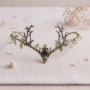 May include: A bronze-toned metal headpiece featuring a central deer head motif with a black faceted stone, flanked by branching antlers and delicate light green leaves attached to a thin wire band that curves to fit around the head.