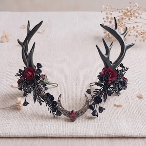 May include: A headpiece featuring two black deer antlers, dark red roses, and black foliage. The centre displays a metallic crescent moon charm with an embedded marquise-cut red gemstone. The structure is connected by a thin, flexible metal band.