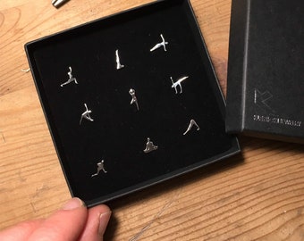Puede incluir: Una caja negra con interior de terciopelo negro que contiene nueve pendientes de plata con poses de yoga. La caja tiene el texto "KLANDISH JEWELRY" impreso en el lateral.