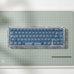 May include: A compact mechanical keyboard featuring a transparent, clear plastic chassis and matching translucent blue keycaps with white lettering. The keyboard layout includes standard QWERTY keys, a spacebar, and modifier keys.