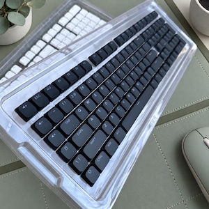May include: A computer keyboard featuring a transparent, clear plastic casing and matte black keys with white lettering. The layout includes standard QWERTY keys, function keys, and dedicated media control keys on the left side.