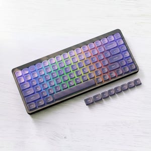 May include: A mechanical keyboard featuring a black frame and translucent purple keycaps with a vibrant rainbow-coloured backlight shining through. A separate row of eight additional purple keycaps is positioned below the main keyboard unit.