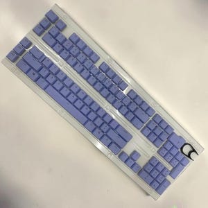 May include: A mechanical computer keyboard featuring a translucent white frame and light purple keycaps. The layout includes a full numeric keypad, function keys, and five dedicated macro keys on the left side. A small black keycap puller is included.