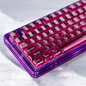 Peut inclure: Un clavier mécanique doté d'un cadre en acrylique violet profond translucide et de touches transparentes assorties avec des lettres blanches. La disposition QWERTY comprend des touches de fonction, une barre d'espace et des touches de modification.