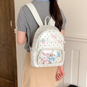 Könnte beinhalten: Ein weißer Rucksack mit schwarzem Punktemuster und einer durchsichtigen Vordertasche. Die Tasche ist mit Hello Kitty-Aufnähern verziert, darunter eine Erdbeere und eine Schleife, und enthält zwei Hello Kitty-Plüschfiguren im Sichtfach.