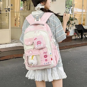 Puede incluir: Una mochila de color rosa claro y crema que presenta un pequeño estuche rectangular con una ilustración de un gato rosa y la palabra "picture" impresa. La bolsa incluye un bolsillo frontal transparente con un peluche blanco y botones rosas.