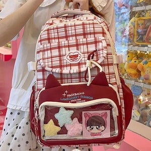 Könnte beinhalten: Ein rot-weiß karierter Rucksack mit einer dunkelroten Vordertasche mit Katzenohren, einem Sichtfenster mit drei Plüschsternen und einer Zeichentrick-Karte sowie dem Text PACKBACK LAMSQOORRT FASHION SINCE 1996 GOODTIME.