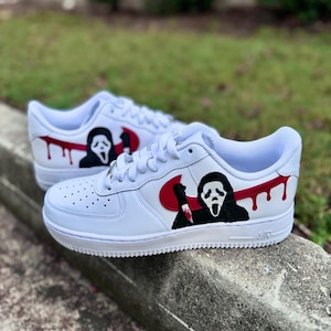 Może przedstawiać: Białe sneakersy z ręcznie malowaną grafiką przedstawiającą postać Ghostface z serii filmów Krzyk. Projekt zawiera czerwone logo swoosh, postać w czarnej szacie trzymającą nóż oraz czerwone krople farby na bocznych panelach.
