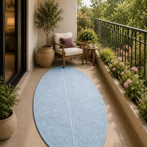May include: An oval-shaped light blue area rug with a textured, embossed floral pattern and a prominent vertical seam running through the centre, placed on a tiled balcony floor in front of a wooden chair and potted plants.