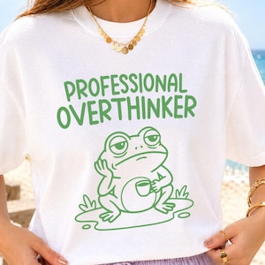 May include: A white short-sleeved t-shirt featuring a green graphic print of a frog sitting and holding a small cup. Above the frog, the text reads "PROFESSIONAL OVERTHINKER" in a bold, green, rounded font. The design is centred on the chest.