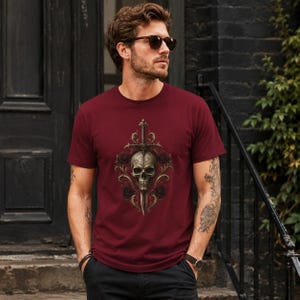 May include: A maroon short-sleeved t-shirt featuring a central graphic print of a human skull pierced by a vertical sword, surrounded by ornate gold filigree scrollwork and dark red roses, with a crew neckline and a standard fit silhouette.