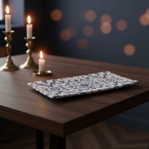 May include: A rectangular ceramic tray featuring a detailed blue and white floral pattern. The tray is positioned on a dark wooden surface alongside three brass candle holders, each containing a lit white candle with a visible flame.