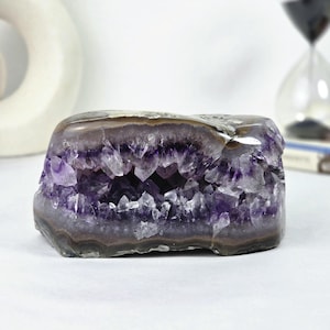 May include: A horizontal amethyst geode featuring a central cavity filled with deep purple, jagged crystal points. The exterior of the stone displays smooth, layered bands of grey and brown mineral deposits, creating a natural contrast.