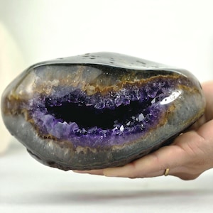 May include: A polished amethyst geode with a smooth, dark brown and black outer shell. The centre features a deep purple, crystalline cavity with jagged, sparkling points. The stone has a rounded, organic shape with a distinct, layered texture.