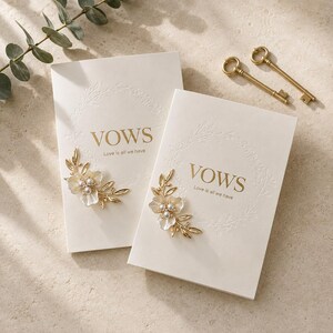 May include: Two cream-colored vow booklets featuring the gold embossed text VOWS and Love is all we have, surrounded by a delicate embossed wreath. Each booklet is adorned with a gold-toned floral embellishment with white pearl centers.