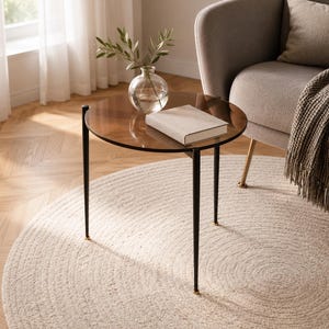 May include: A round side table with a dark brown tinted glass top and three slender black metal legs. A clear glass vase containing green leafy branches and a closed beige hardcover book are placed on the surface of the table.