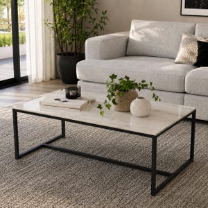 Pode incluir: Uma mesa de centro retangular com tampo de mármore branco e estrutura de metal preto. Sobre a mesa há uma pilha de livros, um pequeno castiçal de vidro preto, uma planta verde num vaso bege e um vaso de cerâmica branco liso.