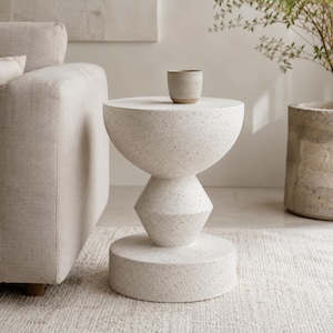 Peut inclure: Une table d'appoint blanche avec une finition mouchetée et une base géométrique sculpturale empilée, composée d'un plateau circulaire, d'une section centrale hexagonale et d'une large base cylindrique. Une petite tasse en céramique assortie est posée dessus.