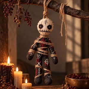 May include: A fabric doll with a cream-colored head featuring black stitched eyes and a mouth, suspended by a jute rope. The body is black with red and white geometric symbols and is wrapped in jute twine, hanging from a dark wooden branch.