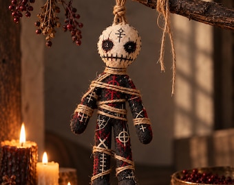May include: A fabric doll with a cream-coloured head featuring black stitched eyes and a mouth, suspended by a jute rope. The body is black with red and white geometric symbols and is wrapped in jute twine, hanging from a dark wooden branch.