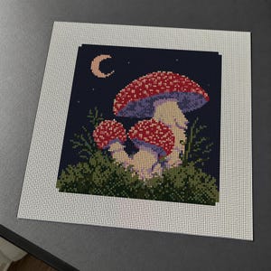 May include: A square cross-stitch pattern on white fabric featuring a dark blue night sky with a crescent moon and stars. The design depicts two red and white spotted mushrooms nestled in green mossy foliage, rendered in a pixelated grid style.