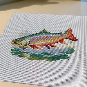 May include: A square piece of white needlepoint canvas featuring a cross-stitched design of a fish. The fish is rendered in shades of green, yellow, orange, and red, depicted swimming above dark green water with visible individual stitches.