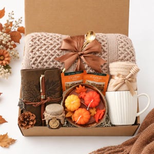 Peut inclure: Un coffret cadeau contenant une couverture en tricot beige avec un ruban marron, un journal marron foncé gaufré de feuilles, un stylo en bois, deux sachets de thé Ceylan, un petit pot avec une étiquette merci, des bougies citrouilles et un mug blanc.