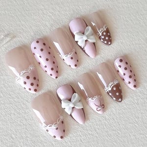Puede incluir: Un juego de uñas postizas en forma de almendra con acabado brillante en una paleta de colores rosa claro y marrón. Los diseños incluyen lunares marrones sobre base rosa claro, detalles de encaje blanco y lazos blancos tridimensionales.