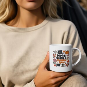 May include: A white ceramic mug featuring a graphic print with the text "Deploy, Debug & Pumpkin Spice" alongside illustrations of a laptop, a pumpkin, autumn leaves, a coffee cup, a flower, and coding symbols in orange, brown, and black colors.