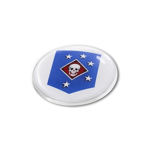 May include: A circular, clear glass cabochon featuring a blue shield-shaped emblem. The emblem displays a central red diamond containing a white skull, surrounded by five white stars arranged in an arc on the blue background of the shield.