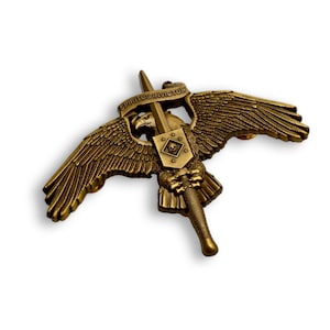 May include: A bronze-toned metal pin featuring an eagle with outstretched wings, clutching a vertical sword. A banner above the eagle reads SPIRITUS INVICTUS. A central shield displays a skull within a diamond shape surrounded by four small stars.