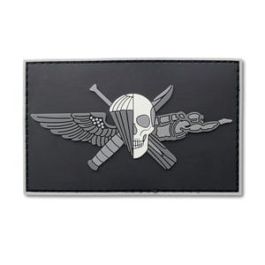 May include: A rectangular black morale patch featuring a central graphic of a skull split vertically, with one side showing a parachute canopy and the other a winged figure. Crossed behind the skull are a knife and a baton in grey tones.