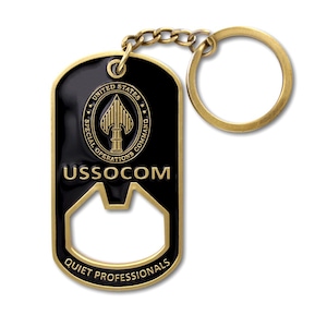 May include: A black and gold metal keychain featuring a bottle opener design. The tag displays the United States Special Operations Command emblem, the text USSOCOM, and the phrase QUIET PROFESSIONALS in gold lettering against a black background.