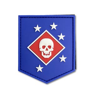 May include: A shield-shaped morale patch in blue, featuring a central red diamond with a white skull emblem. Five white stars are arranged around the diamond on the blue background. The patch has a stitched border and a flat, molded surface design.