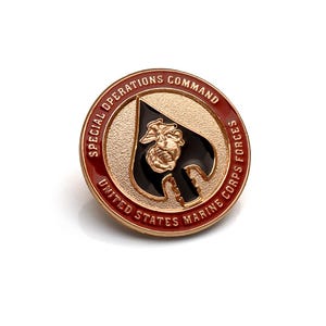 May include: A circular gold-tone metal pin featuring a black spade emblem in the center with a gold eagle, globe, and anchor insignia. The outer red border contains the embossed text SPECIAL OPERATIONS COMMAND UNITED STATES MARINE CORPS.