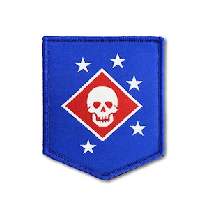 May include: A blue embroidered patch with a shield-like shape featuring a central red diamond containing a white skull. Five white stars are arranged in an arc around the diamond on the blue background, with a stitched border around the edge.