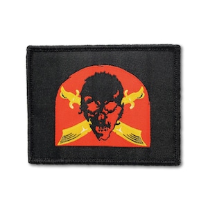 Könnte beinhalten: Ein rechteckiger, schwarz bestickter Aufnäher mit einem zentralen roten, halbkreisförmigen Emblem. Das Design zeigt eine schwarze Totenkopf-Silhouette über zwei gekreuzten gelben Dolchen mit detaillierten Griffen und Klingen auf schwarzem Grund.