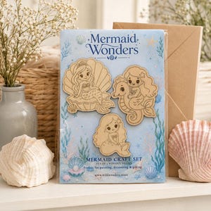 Könnte beinhalten: Ein Set aus drei hellen Holzfiguren in Meerjungfrauenform, verpackt auf einer blauen Karte mit dem Text Mermaid Wonders. Die Formen zeigen eine Meerjungfrau in einer Muschel, eine Meerjungfrau auf einem Seepferdchen und eine sitzende Meerjungfrau.