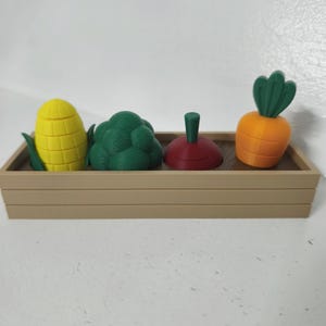 Può includere: Una fioriera rettangolare color marrone chiaro contenente quattro piccole figure di verdure in plastica: una pannocchia di mais gialla, un broccolo verde, un pomodoro rosso scuro con gambo verde e una carota arancione con foglie verdi.