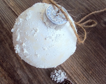 May include: A white, textured Christmas ornament with a silver cap and twine string, resting on a rustic wooden surface. Two small pinecones are nearby. The ornament has a frosted appearance, suggesting a winter theme.