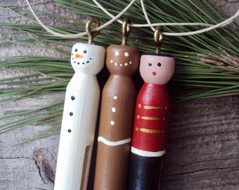 Könnte beinhalten: Drei hölzerne Weihnachtsschmuckstücke: ein Schneemann, ein Lebkuchenmann und ein Spielzeugsoldat. Der Schneemann ist weiß mit schwarzem Zylinder und Kohleaugen. Der Lebkuchenmann ist braun mit weißem Zuckerguss. Der Spielzeugsoldat hat einen roten Mantel und eine schwarze Hose.